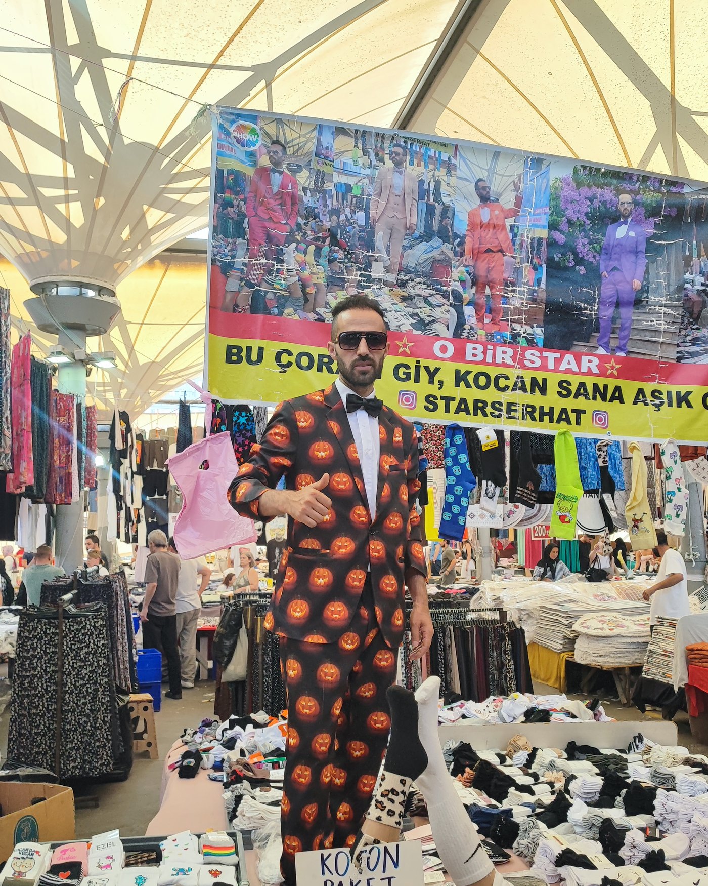 Star Serhat'ın çorap tezgâhı — arkasında kendi afişi, önünde paket paket çoraplar